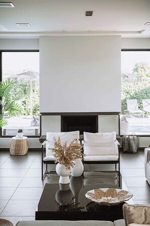 A modern living room with a fireplace, two white sofas, a coffee table, and a view of greenery outside. - Villa Vega Heated* pool, next to nature! (Chania)