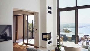 A modern living room with a large window, a sofa, a dining table, and a TV. - Omnia Villa l - Heated* pool & stunning Seaview! (Chania)