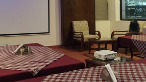 A room with a large projection screen, a projector, and a table with a red tablecloth. There are two chairs and a bed with a red and white checkered bedspread. - Hotel Eurasia Mohali Airport (Mohali)