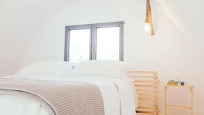 A bedroom with a bed, a window, a bedside table with books, and a wall-mounted light fixture. - Exi Sea Side Suites (Santorini)