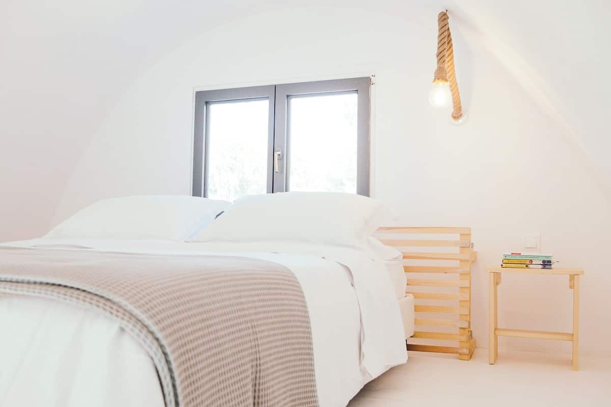 A bedroom with a bed, a window, a bedside table with books, and a wall-mounted light fixture.
