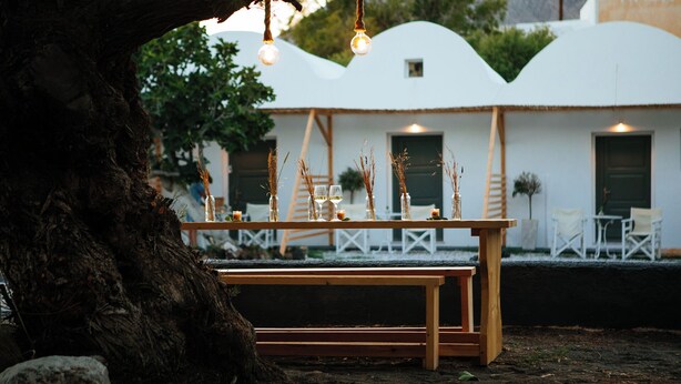 A wooden bench with a table set for a meal, surrounded by trees and a white building with a unique architectural design in the background. - Exi Sea Side Suites (Santorini)