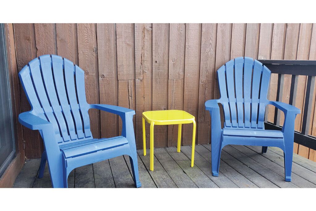 Two Adirondack chairs and a small yellow table on a wooden deck.