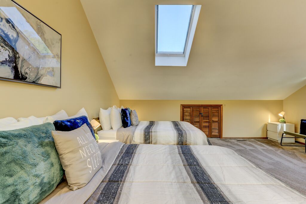 A bedroom with a skylight, two beds, a painting, and a desk with a lamp.