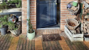 Un edificio de ladrillo con techo de tejas, una puerta azul oscuro y un pequeño jardín con plantas en macetas y un banco de madera.