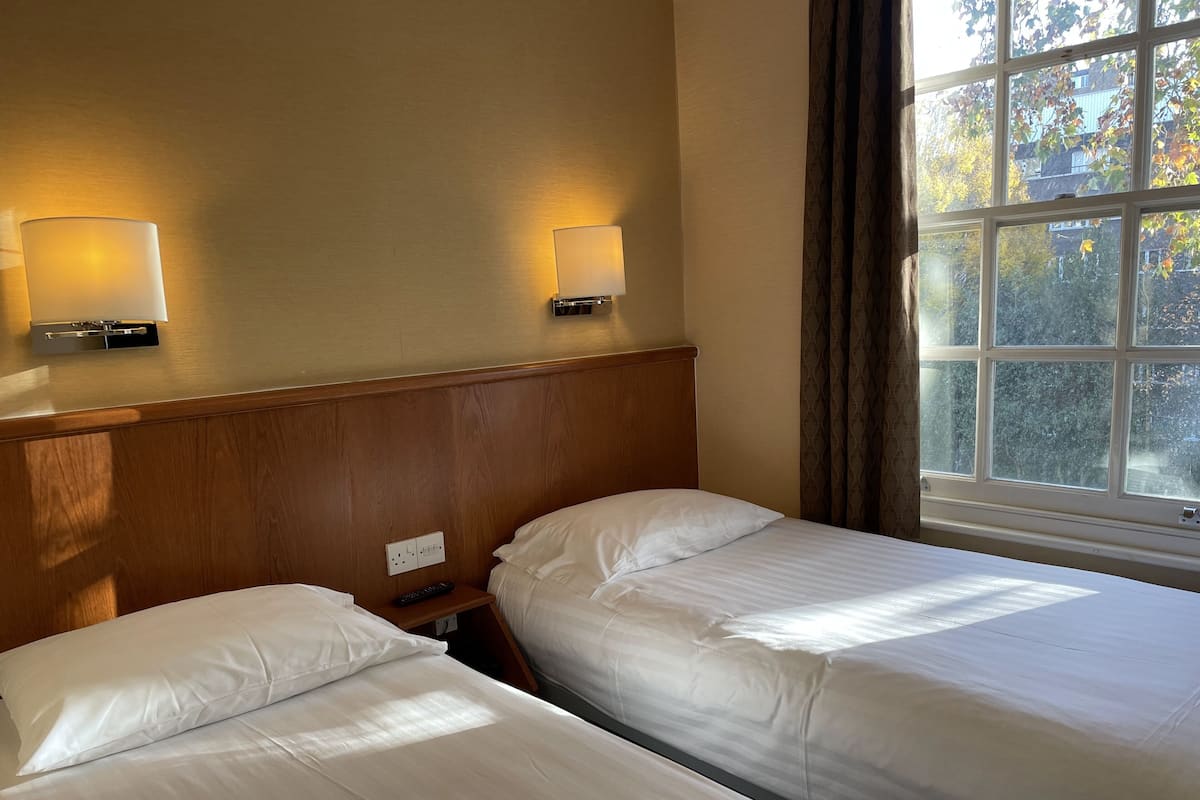 A hotel room with two beds, a wooden headboard, bedside lamps, a window with a view of trees, and a power outlet on the wall.