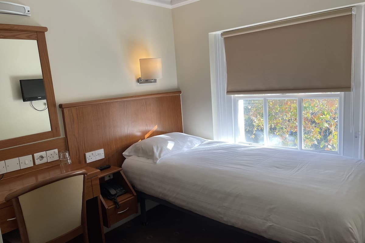 A hotel room with a bed, a desk with a chair, a telephone, a mirror, and a window with a view of trees.