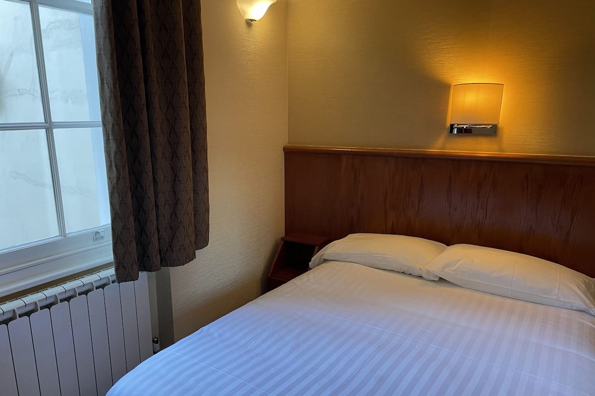 A hotel room with a bed, a window with curtains, a wall-mounted light, and a radiator.