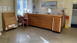 Un espace d’accueil comprenant un comptoir de réception en bois, une chaise, un présentoir de brochures, une fenêtre donnant sur des bâtiments et un panneau d’avertissement au sol.