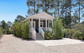 A small, single-story house with a porch, surrounded by trees and gravel driveway.