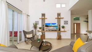 Ein modernes Wohnzimmer mit einer Couch, einem Couchtisch und einem an der Wand befestigten Fernseher. Eine Glastür ermöglicht den Zugang zu einem Balkon.