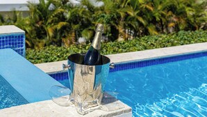 Eine Flasche Champagner in einem Metallkübel neben einem Pool mit blauen Kacheln und tropischen Pflanzen im Hintergrund.