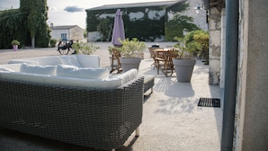 A wicker sofa with cushions, a table, chairs, and a purple umbrella in a courtyard with a building in the background.