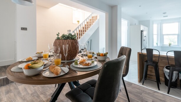 Un comedor moderno con una mesa de madera dispuesta para el desayuno, que incluye cruasanes, fruta y jugo de naranja.