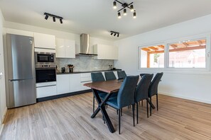 Eine moderne Küche mit Edelstahlgeräten, ein Essbereich mit Holztisch und Stühlen sowie ein großes Fenster mit Blick ins Freie.