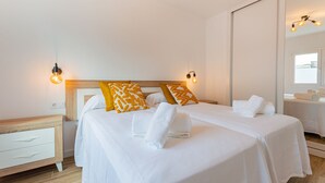 Un lit double avec une literie blanche, deux oreillers de couleur moutarde et une tête de lit en bois. Il y a deux tables de chevet avec des tiroirs blancs et un luminaire fixé au mur.