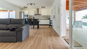 Un salon moderne avec un canapé gris, un coin repas avec des chaises, une cuisine avec des meubles blancs et une terrasse en bois visible à travers des portes vitrées.