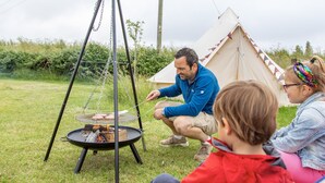 Mies grillaa ulkona kannettavan grillin avulla. Taustalla on teltta ja edessä ruohikkoalue.