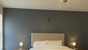 Une chambre à coucher avec une tête de lit capitonnée, un ventilateur de plafond, deux lampes de chevet et une fenêtre avec des stores.