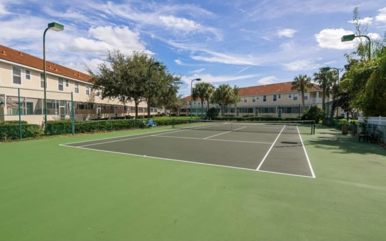 A tennis court with a green surface, surrounded by a fence, and adjacent to a building with a red roof.
