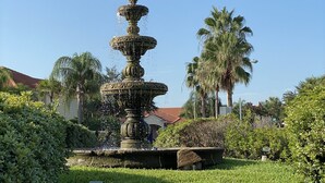 Une fontaine à plusieurs niveaux, située dans un jardin bien entretenu, agrémenté de palmiers et surplombant un ciel d’un bleu éclatant.