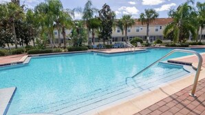 Une piscine rectangulaire bordée d’une terrasse carrelée, entourée de palmiers et avec un bâtiment doté de nombreuses fenêtres en arrière-plan.