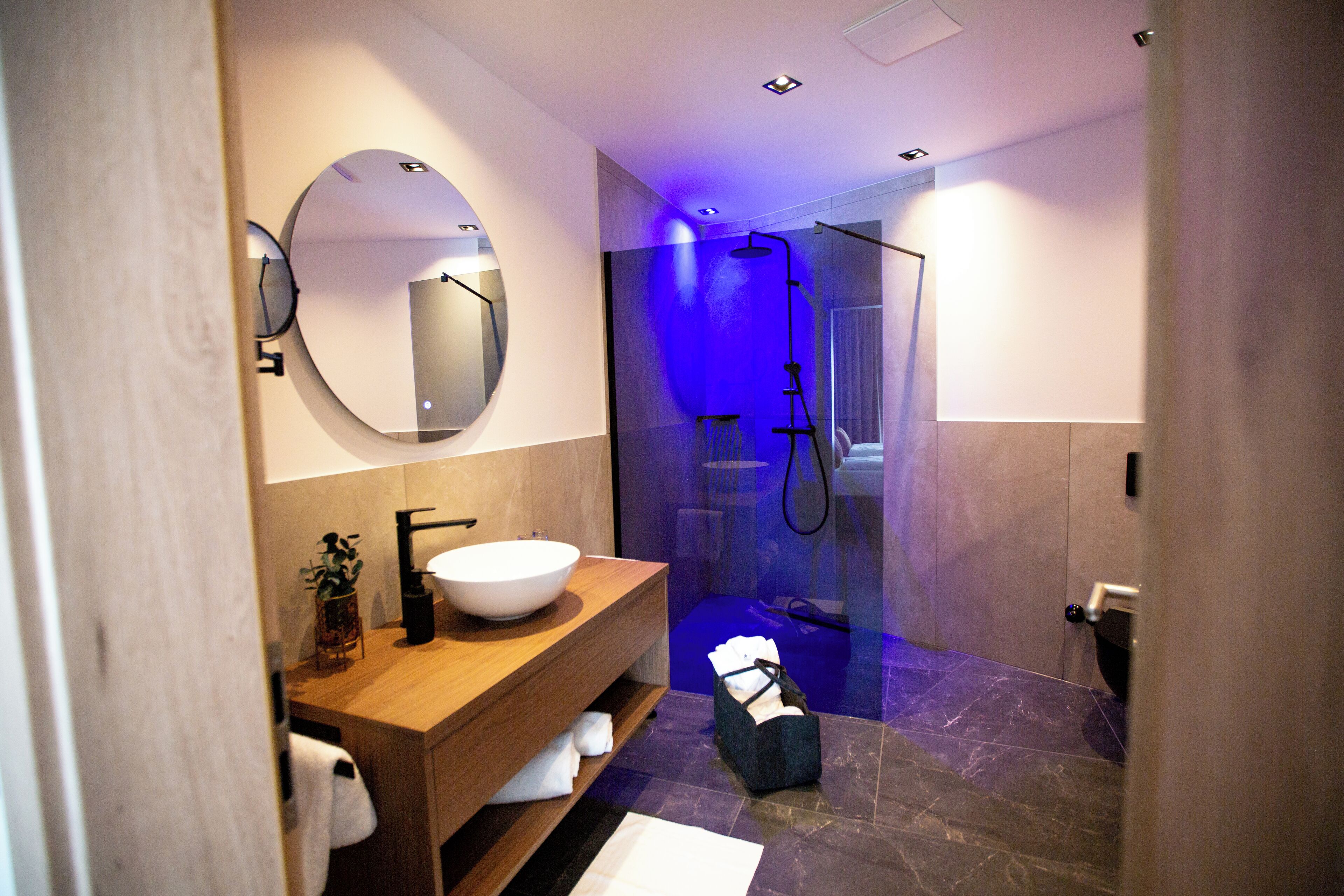 A modern bathroom with a round mirror, wooden vanity, white sink, and a glass-enclosed shower.