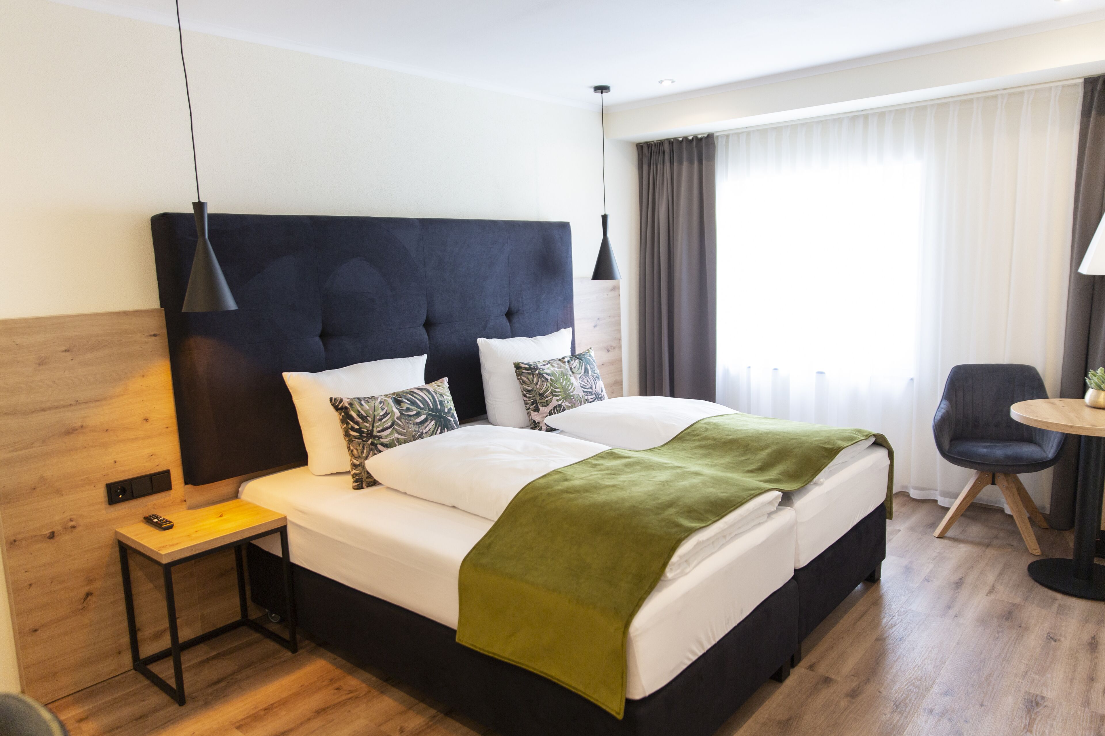 Una habitación de hotel moderna con una cama grande, una mesita de noche, una silla y un ventanal con cortinas.