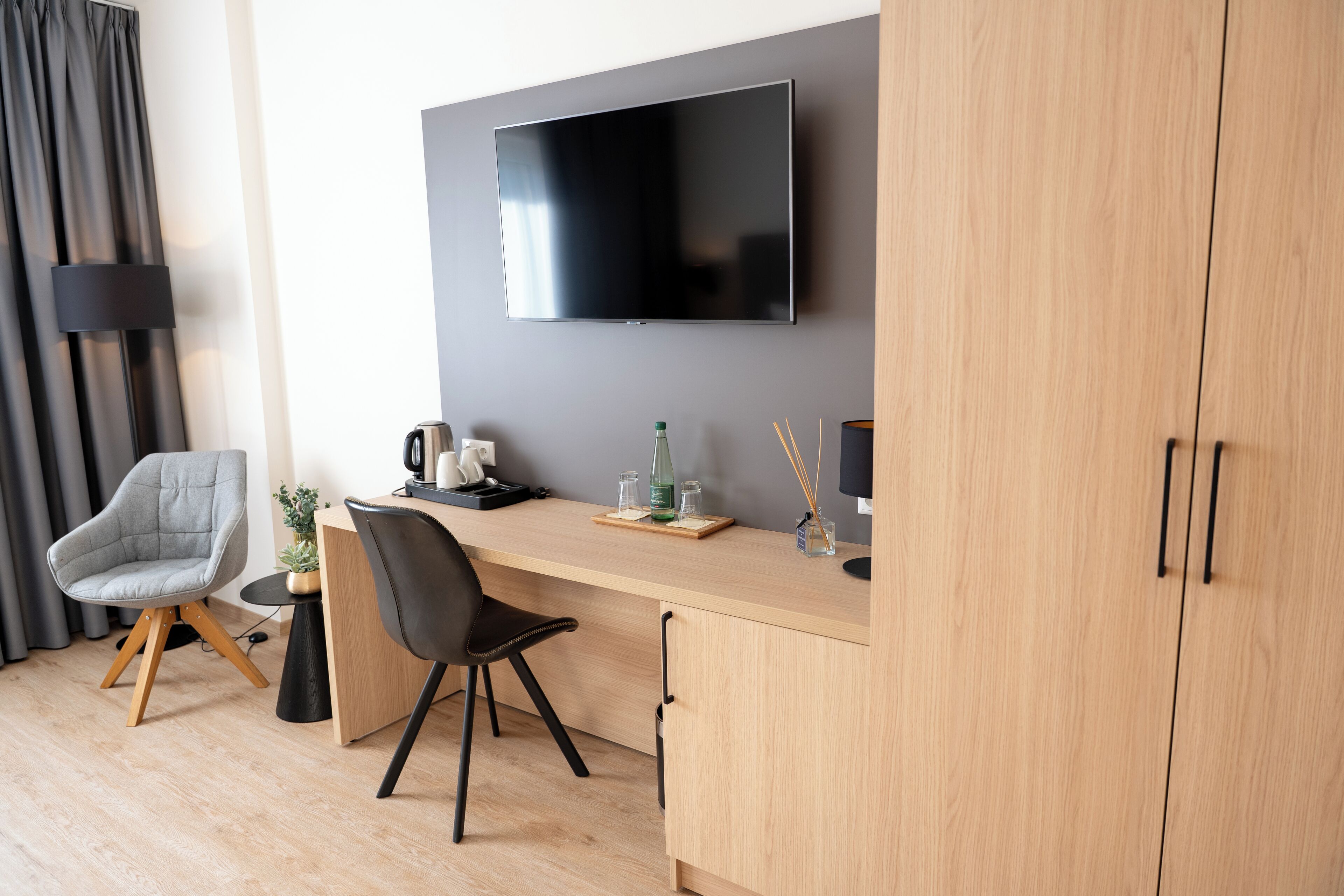 A modern hotel room with a wooden desk, two chairs, a flat-screen TV mounted on the wall, a small side table with a plant, a coffee maker, and a glass of water.