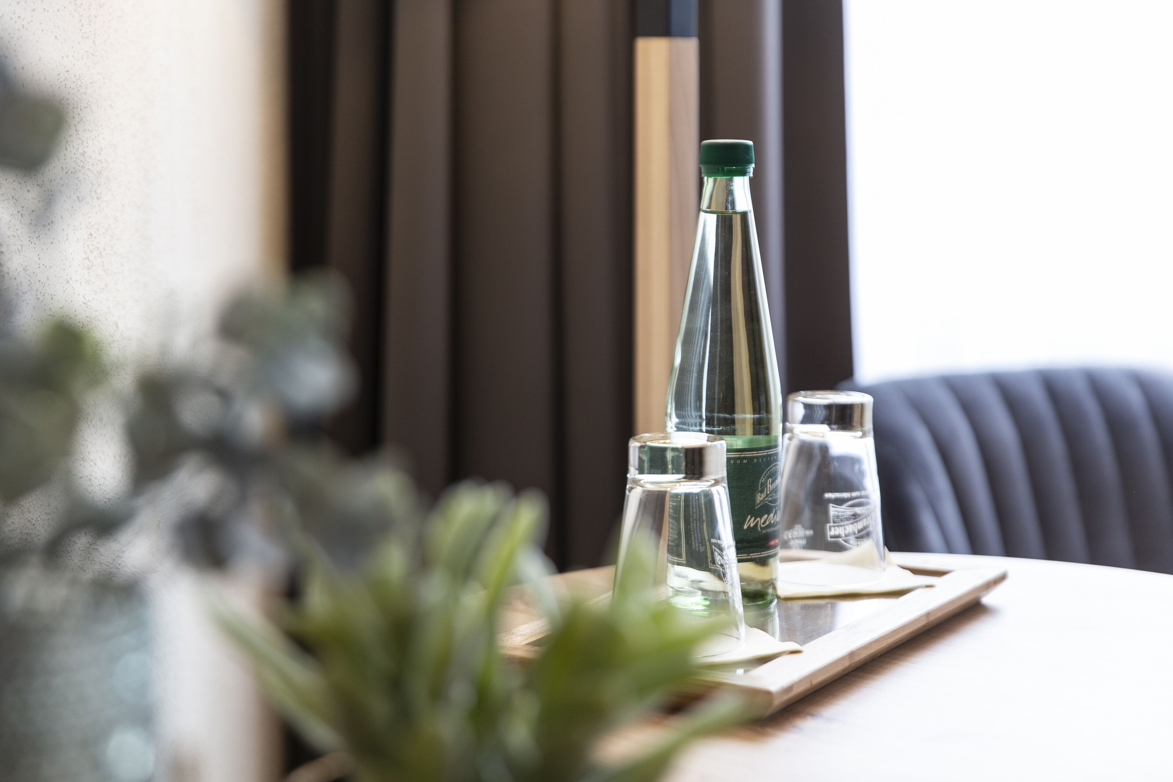Une table avec une bouteille et deux verres, une plante en pot et un fauteuil à l’assise sombre.