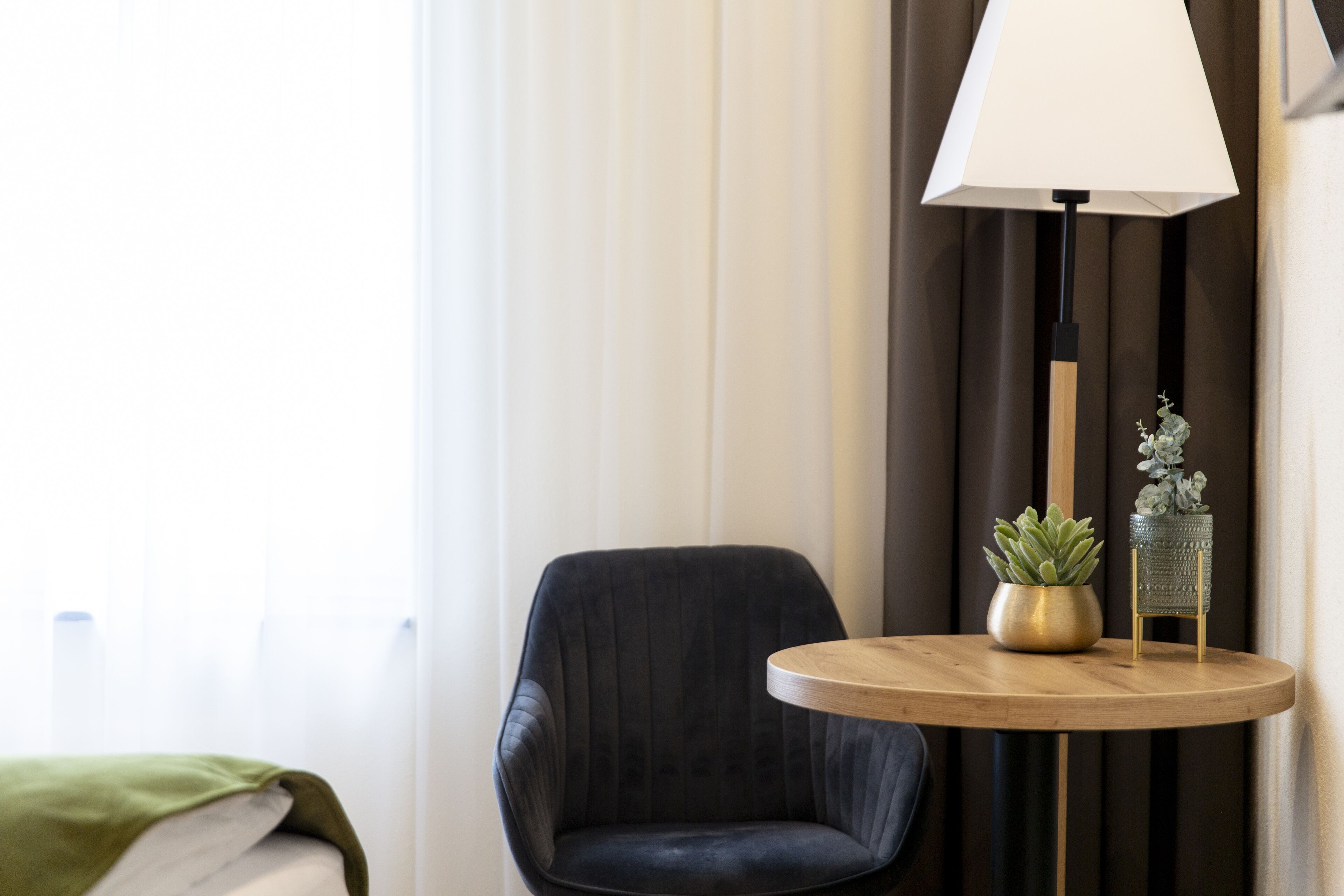 Een moderne hotelkamer met een donker beklede fauteuil, een rond houten bijzettafeltje met een lamp en een potplant.