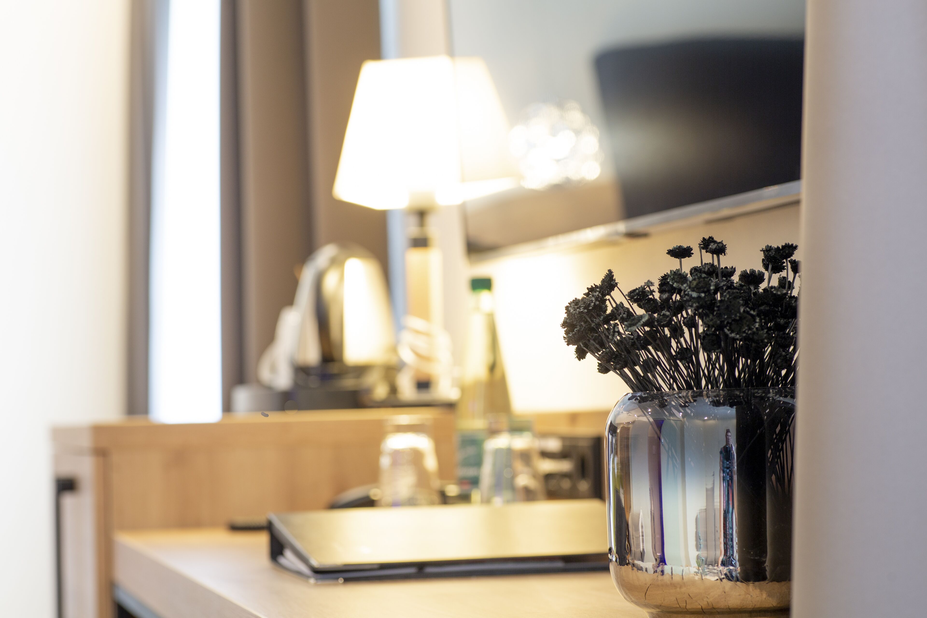A hotel room with a bedside table, a lamp, a laptop, and a vase with dried flowers.