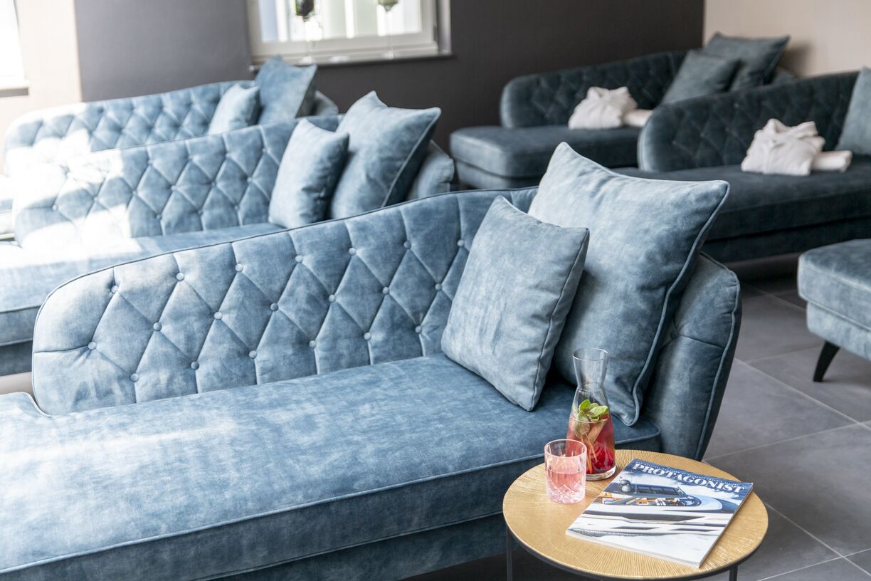 A modern lounge area with tufted blue sofas and armchairs, a round wooden coffee table with a magazine, and a glass of drink.