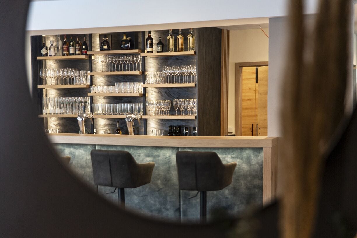 Una zona de bar con estantes llenos de botellas y vasos, dos taburetes y una puerta de madera al fondo.