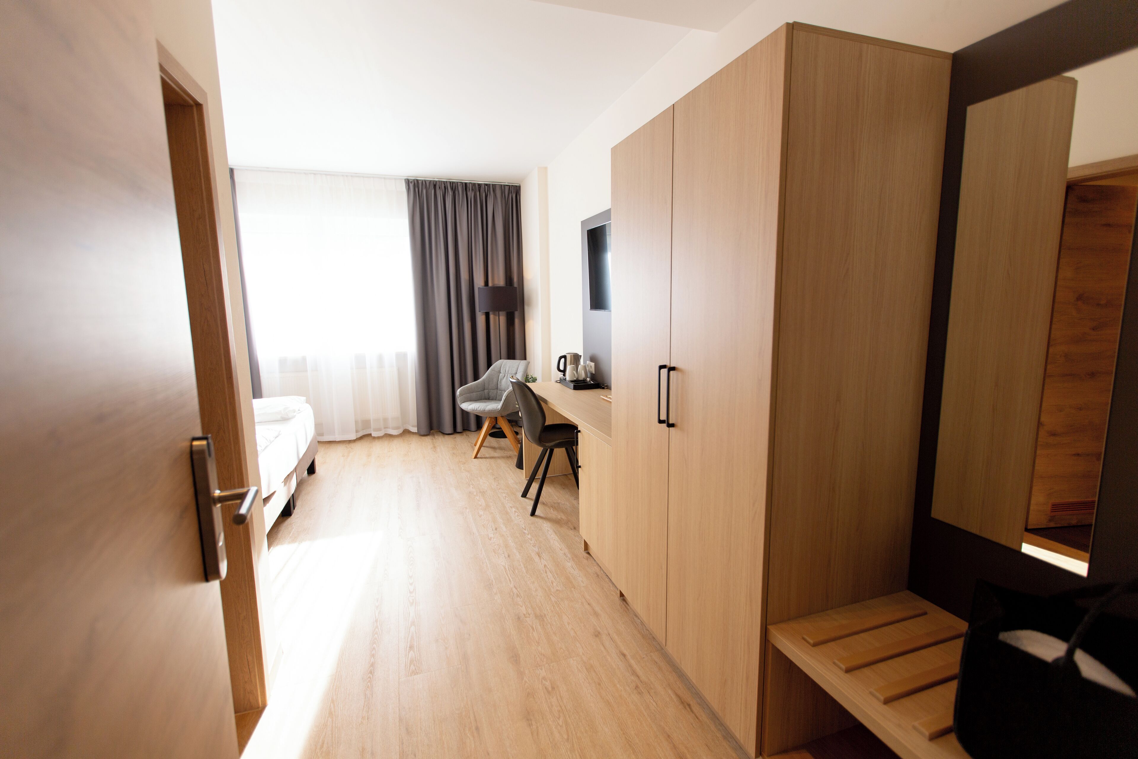 Una habitación de hotel moderna con suelo de madera, una cama, un escritorio con silla, un armario y un gran ventanal con cortinas.