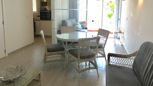 A modern living room with a glass-top dining table surrounded by wicker chairs, a grey sofa, and a small side table with a candle.