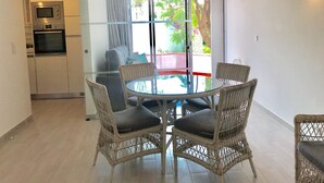 A modern kitchen with a built-in microwave and oven, a dining area with wicker chairs and a glass table, and a sliding glass door leading to a balcony with a view of greenery.