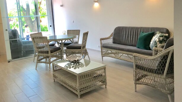 A modern living room with wicker furniture, a glass coffee table, and a grey sofa with a green pillow.