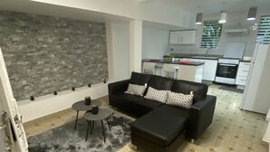 Ein modernes Wohnzimmer mit einem schwarzen Sofa, einem Couchtisch und einem gemusterten Teppich. Im Hintergrund ist eine Küche mit weißen Schränken und einer gefliesten Spritzschutzwand zu sehen.