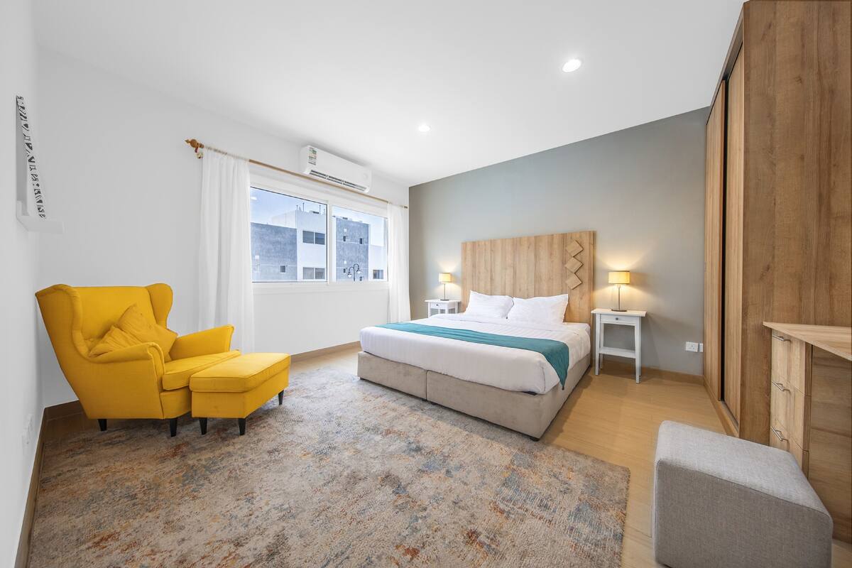 A modern bedroom with a bed, a yellow armchair, a small table with a lamp, and a wooden wardrobe.