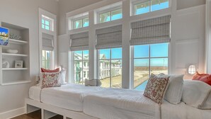A room with a large window seat, white walls, and a view of a building and beach.