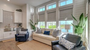 A spacious living area with a beige sofa, two armchairs, and a coffee table. There are large windows with blinds, a kitchen with white cabinets, and a view of a green landscape.