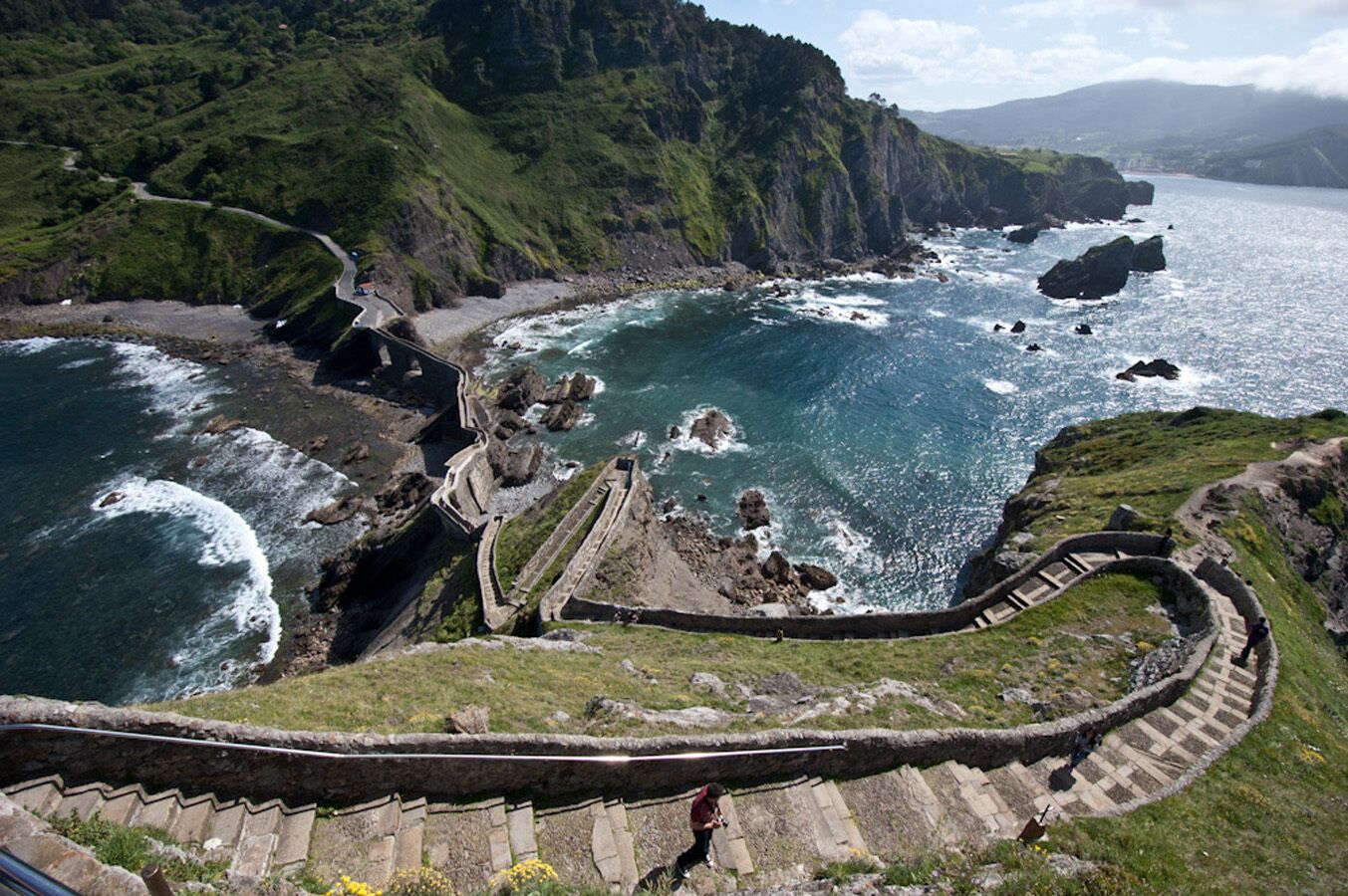 Un paisaje costero con una escalera de piedra que desciende hacia el mar, rodeado de acantilados y vegetación.