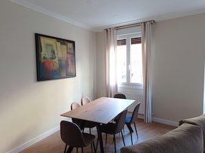 A dining area with a wooden table and chairs, a painting on the wall, and a window with curtains.
