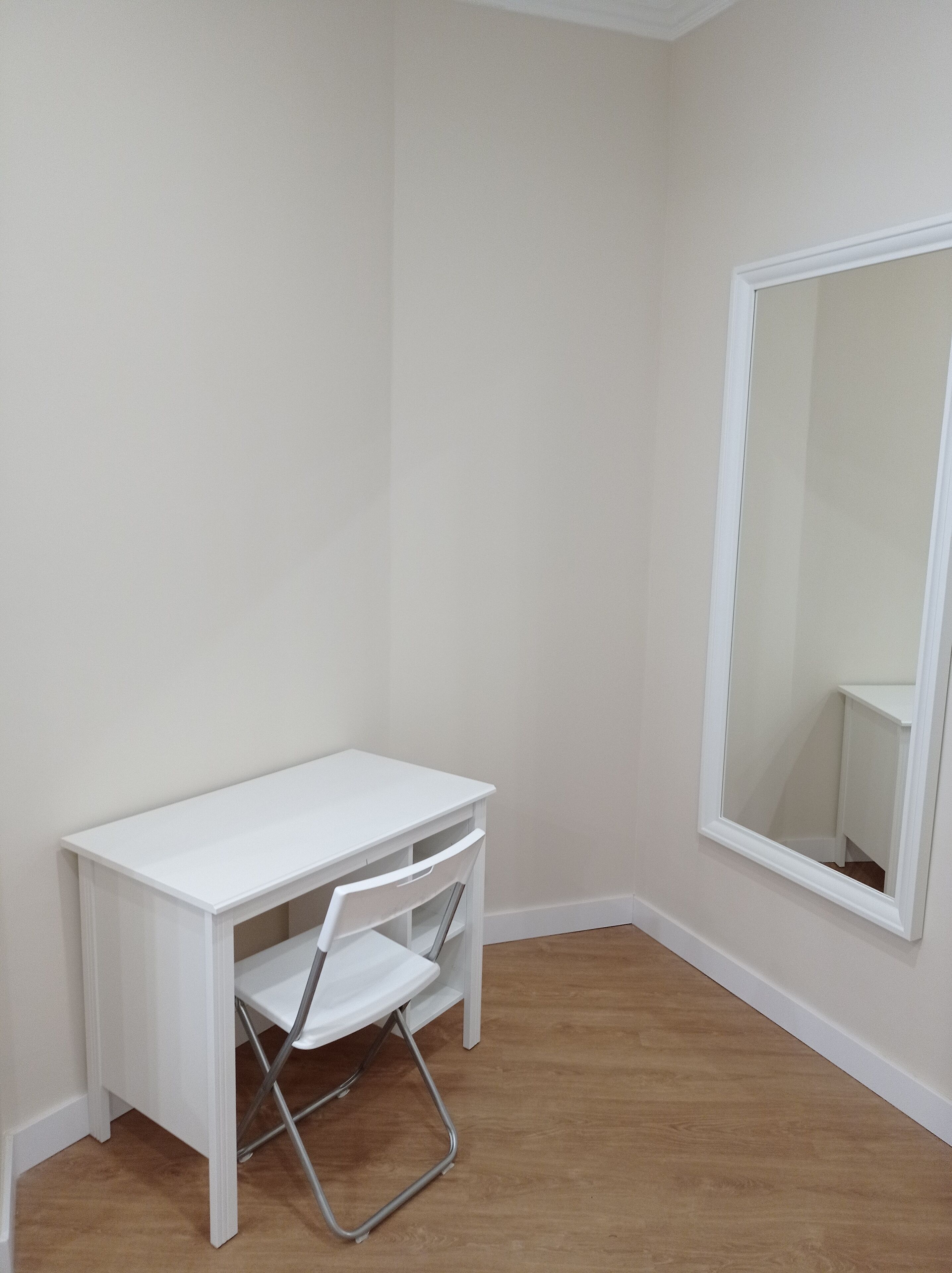 Un escritorio pequeño y blanco con una silla en una habitación de esquina, con suelo de madera y un espejo grande en la pared.