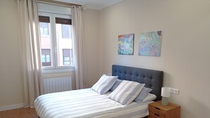 A bedroom with a bed, a nightstand, a window with curtains, and two wall-mounted paintings. - URDAIHOME APARTMENT (Gernika-Lumo)