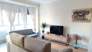 A modern living room with a grey sofa, a flat-screen TV on a wooden console, a potted plant, and a painting on the wall. - URDAIHOME APARTMENT (Gernika-Lumo)