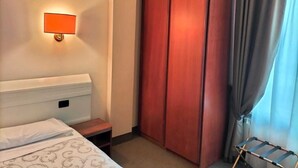 A hotel room with a bed, a wardrobe, a nightstand, a lamp, and a window with curtains.