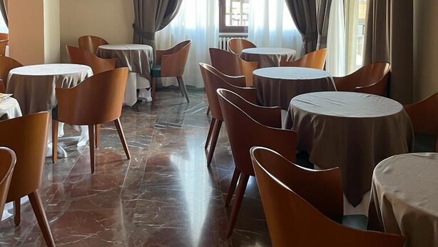 A dining area with round tables and chairs, a flat-screen TV mounted on the wall, and large windows with curtains.