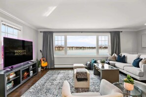 A modern living room with a large TV, a guitar, a sofa, and a view of a body of water. - Waterview House Walking Distance from Cove Beach (Stamford)